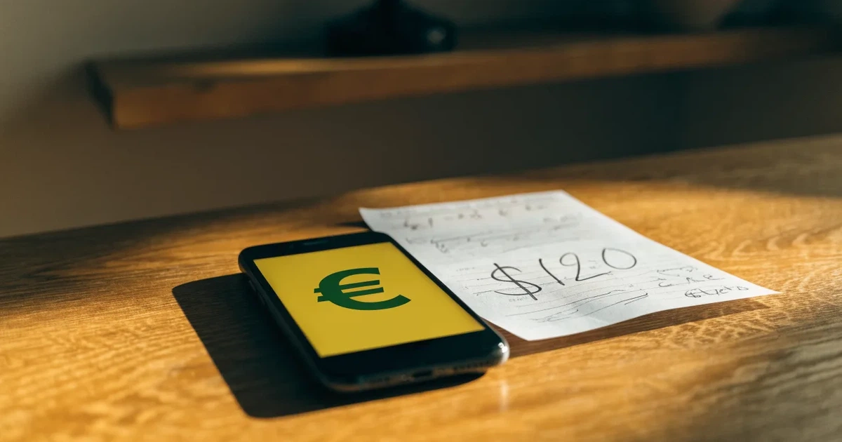Um telemóvel e um cartão bancário lado a lado numa mesa de madeira, com algumas notas de euro ao lado